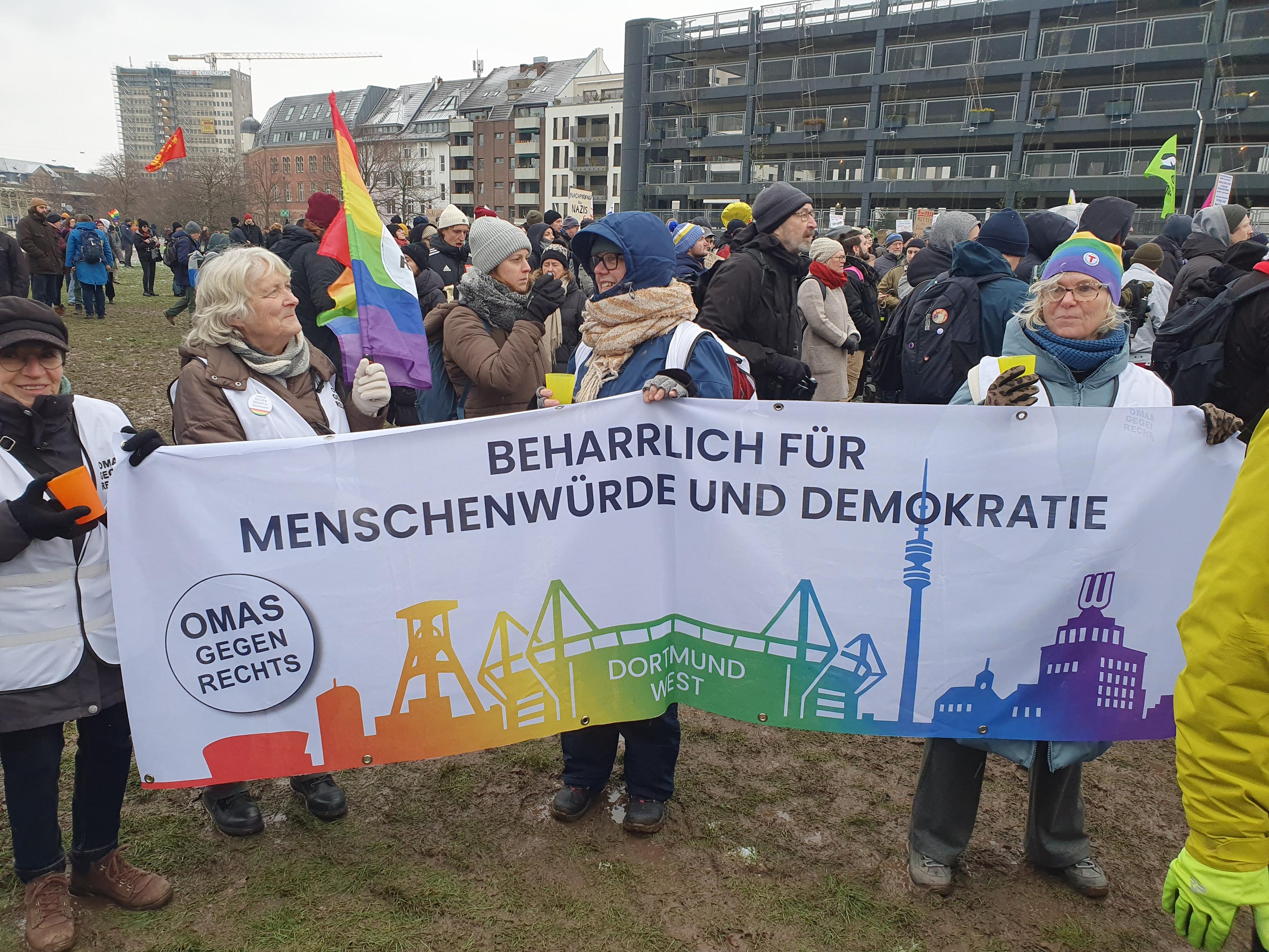 Die Omas aus Dortmund halten ein Banner: einige Wahrzeichen von Dortmund (der Turm, ein Förderturm,das Stadion, usw. als Silhouetten in Regenbogenfarben. Text: BEHARRLICH FÜR MENSCHENWÜRDE UND DEMOKRATIE