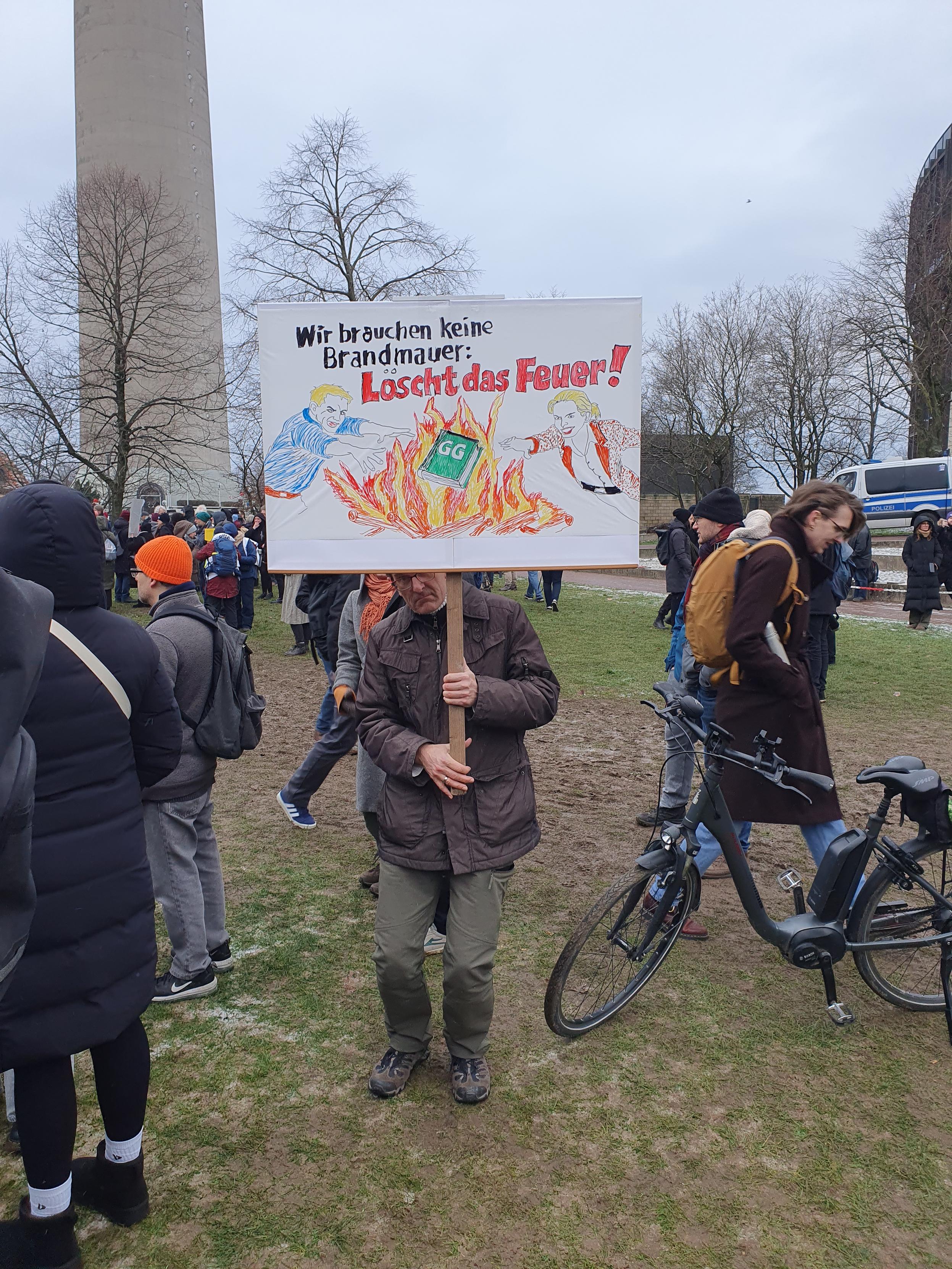 Ein Mensch hält ein Schild mit einem lichterloh brennenden Grundgesetz, Text:wir brauchen keine Brandmauer,löscht das Feuer!