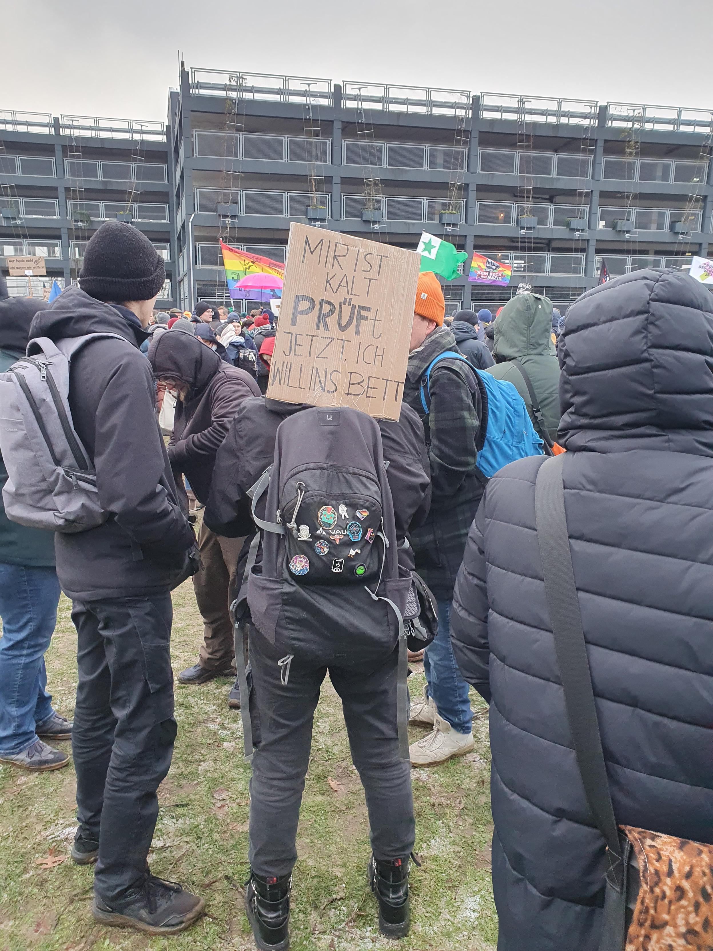 Ein Mensch hat ein Schild am Rucksack befestigt: MIR IST KALT, PRÜF JETZT ,ICH WILL INS BETT