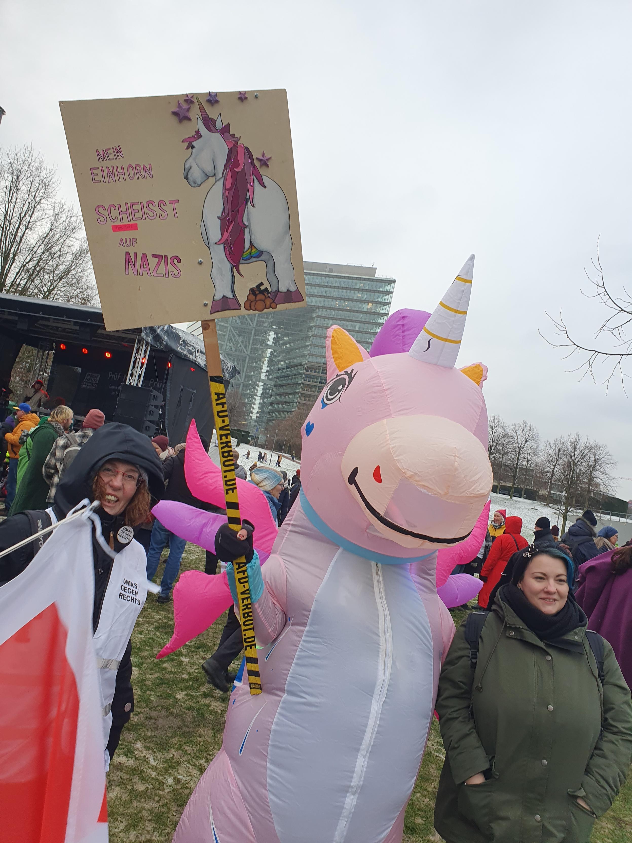 Ein Einhorn trägt ein Schild mit einem Einhorn von hinten, darunter ein Kackhaufen. Text: MEIN EINHORN SCHEISST AUF NAZIS
