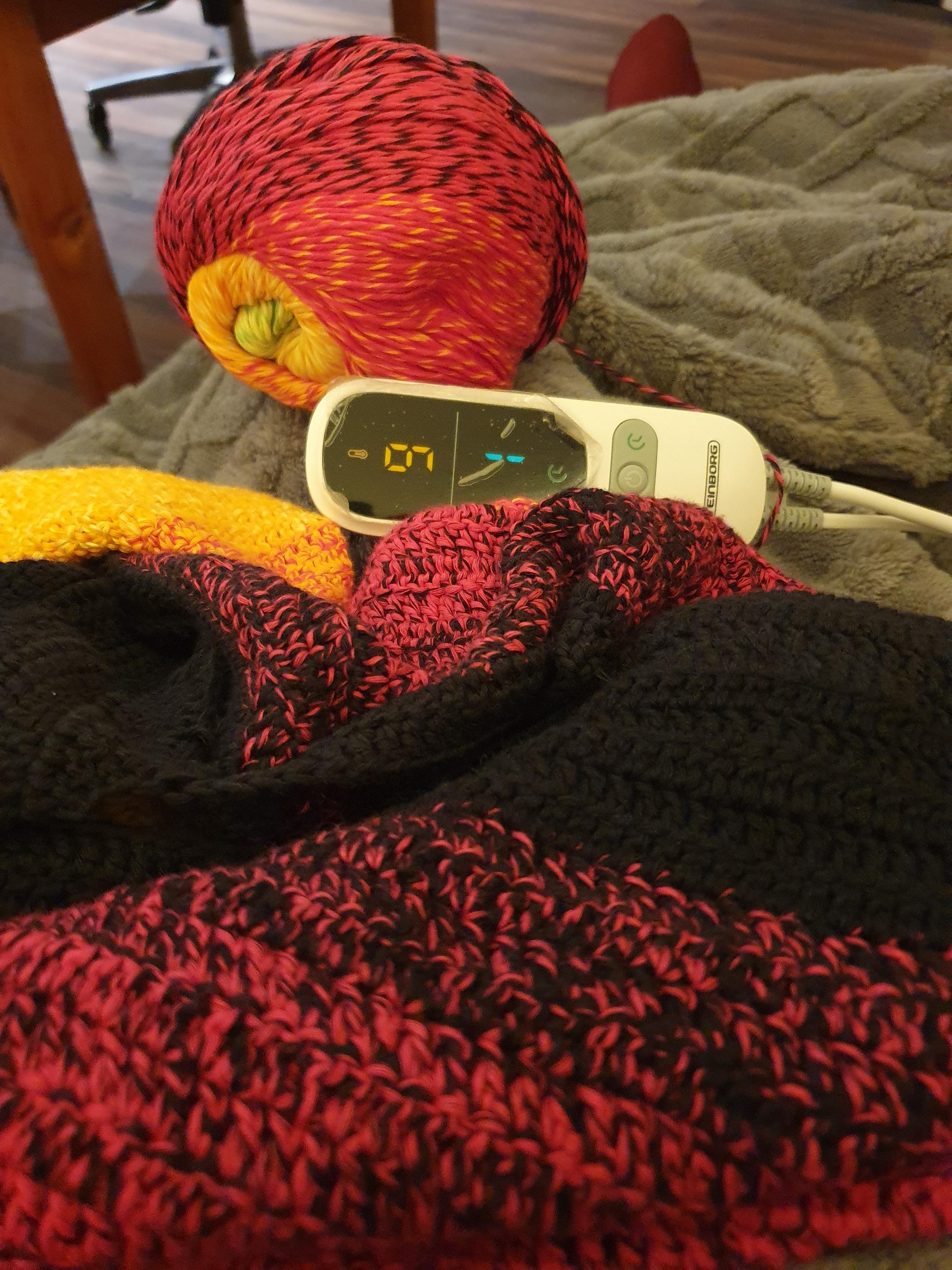 Ein Photo einer Heizdecke auf Stufe 9, darauf ein unfertiger Pullover und Farbverlaufwolle in Regenbogenfarben
